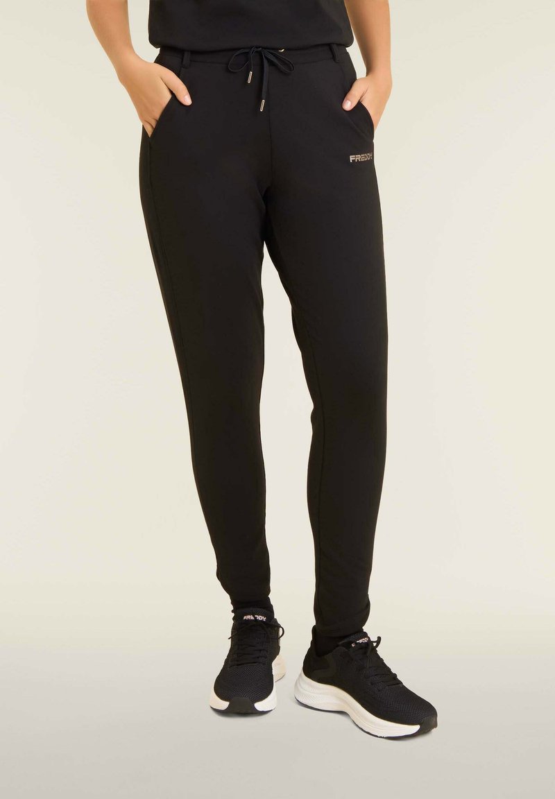 Pantalon de survêtement noir avec une coupe fuselée, taille à cordon, poches latérales et un petit logo doré sur la hanche gauche. Texture lisse.
