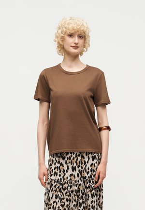 Γυναίκα με κοντά σγουρά ξανθά μαλλιά, φοράει ένα καφέ κοντομάνικο πουκάμισο και μια φούστα με λεοπάρ μοτίβο, στέκεται μπροστά σε απλό φόντο.