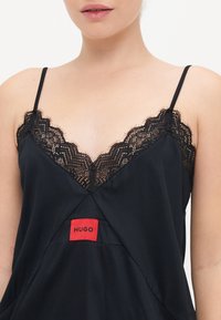 Mustat camisolit, joissa on pitsiyksityiskohtia V-pääntiessä. Ominaisuuksiin kuuluu ohuet olkaimet ja punainen "HUGO" -labeli keskellä. Silkkinen rakenne.
