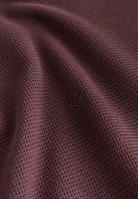 Audinys su tekstūra purpuriniu atspalviu, kuriame yra tinklelio raštas. Medžiaga atrodo minkšta, šiek tiek tampri, su subtiliu logotipo akcentu.