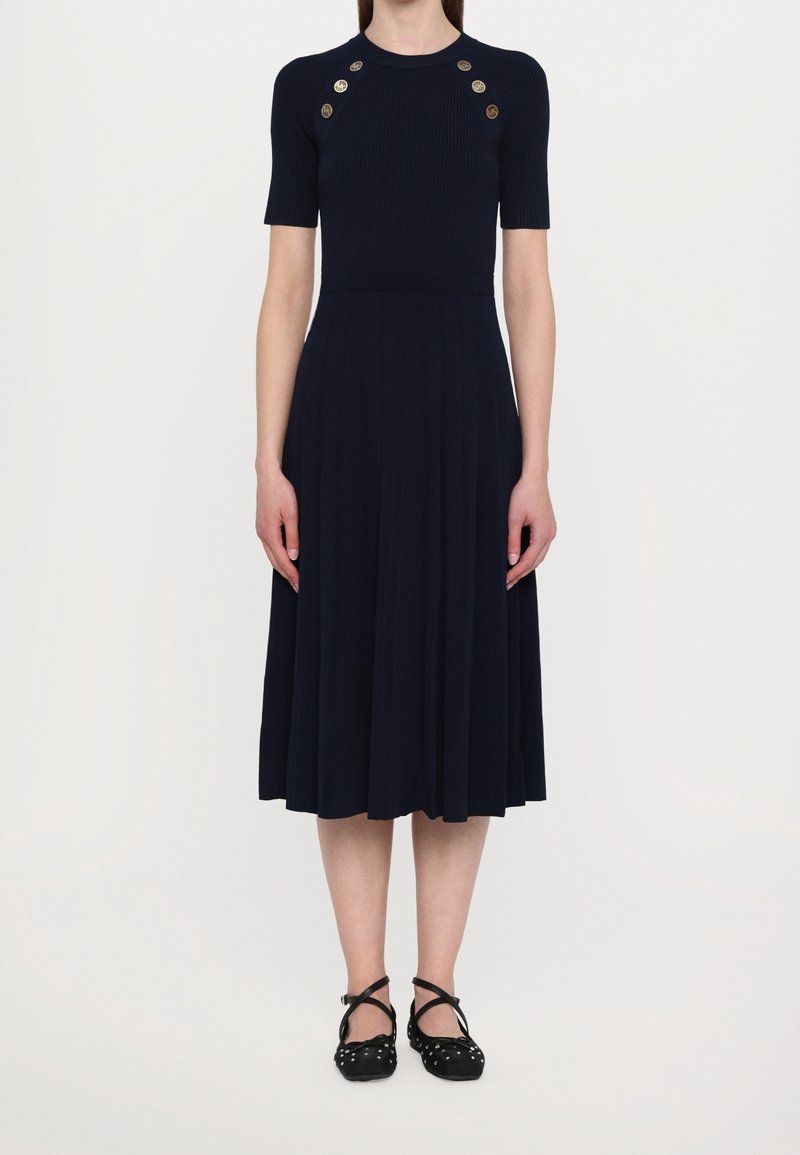 Femme portant une robe midi bleu marine à manches courtes avec des boutons dorés sur la poitrine et des chaussures plates noires avec des brides aux chevilles.