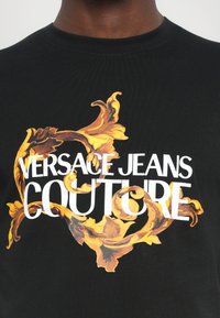 Črna bombažna majica z drznim zlatim cvetličnim vzorcem in belim napisom "VERSACE JEANS COUTURE" na sredini.