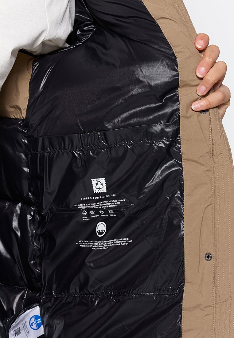 Die beige Pufferjacke verfügt über ein schwarzes Innenfutter, mit aufgedruckten Pflegehinweisen und Logos. Weicher Stoff und isoliertes Design.