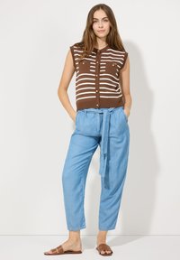 Braun gestreifter ärmelloser Oberteil mit goldenen Knöpfen, kombiniert mit hellblauen, lässigen Hosen und braunen Rutsch-Sandalen.