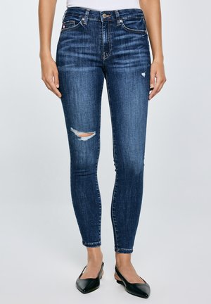 Naine, kes kannab tumesiniseid rebenditega kitsaid teksapükse ja musti teravate ninadega kannavarrega poolkingi, seisab ühevärvilise tausta ees.