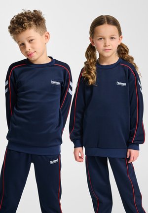 Zwei Kinder tragen identische dunkelblaue Hummel-Trainingsanzüge mit roten und weißen Streifen und stehen vor einem schlichten Hintergrund.