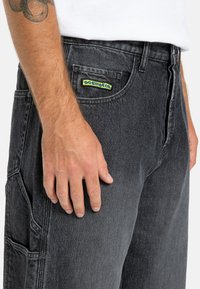 Czarne dżinsowe szorty cargo z żółtą naszywką logo, bocznymi kieszeniami i wytrzymałą strukturą materiału. Dopasowany krój w okolicy talii.