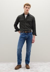 Donkergroen overhemd met knopen en twee borstzakken, blauwe spijkerbroek, bruine gevlochten leren riem en bruine veterschoenen op een neutrale achtergrond.