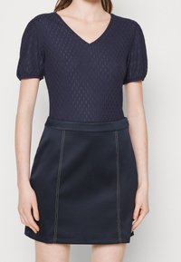 Námořnický top s krátkým rukávem a texturovaným vzorem, s výstřihem do V, v kombinaci s přiléhavou tmavou sukní s bílým prošíváním a hladkým povrchem.