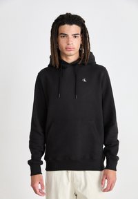 Sudadera con capucha negra fabricada en un tejido suave, con un bolsillo frontal tipo canguro, capucha ajustable con cordón y pequeño logo blanco en el pecho.