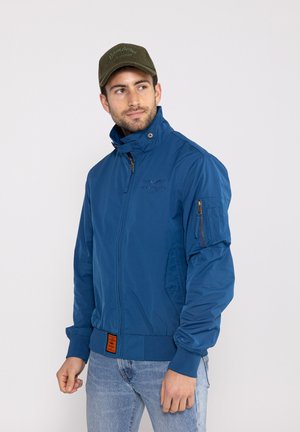 Blaue Bomberjacke mit Reißverschluss vorne, hohem Kragen, seitlichen Taschen und Reißverschlusstasche am Ärmel. Besitzt ein orangefarbenes Etikett an der Taille.