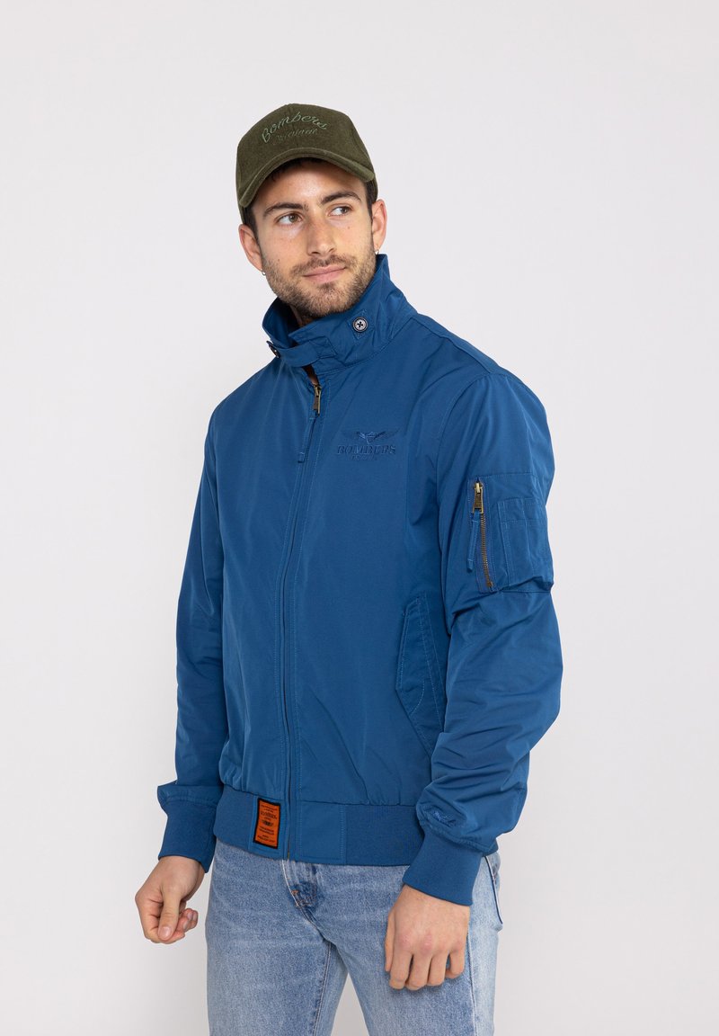 Veste bomber bleue avec fermeture éclair à l'avant, col haut, poches latérales et poche zippée sur la manche. Présente une étiquette orange à la taille.