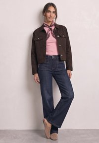 Femme debout portant une veste marron, un top rose avec un foulard à motifs autour du cou, un jean bleu foncé à jambes larges et des chaussures mule beiges.