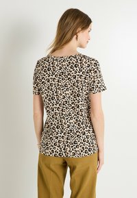 Camiseta de estampado de leopardo en beige y negro con mangas cortas, cuello redondo, tela suave y un corte relajado, vista desde la parte de atrás con pantalones de color claro.