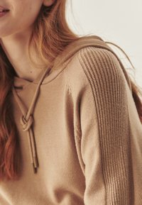 Beżowy hoodie z fakturowanym prążkowanym detalem na ramionach, z kapturem na sznurki i miękkiego materiału. Zbliżenie ukazujące kunszt wykonania.