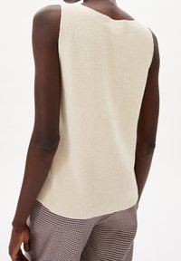 Kermanvärinen hihaton pusero, jossa on teksturoitu neulottu kuvio. Sileä pinta, rento istuvuus ja lievästi kaartuva helma. Päällä ruudulliset housut.