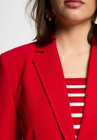 Rode blazer met een gladde textuur en een op maat gemaakte pasvorm, voorzien van schoudernaden. Gelaagd over een rood-wit gestreept top. Gouden oorbellen zichtbaar.
