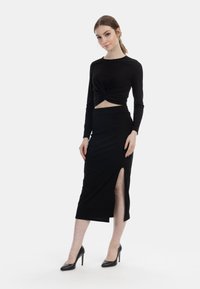 Schwarzes Zweiteiler-Outfit mit einem langärmligen Croptop mit Knotendetail und einem bodenlangen Rock mit hohem Schlitz, kombiniert mit High-Heels.