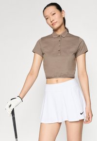 Brūna īsā polo krekls ar pogu detaļām, apvienots ar baltu izplešanos golfa svārku, kurā redzams melns Nike logo. Iekļauta balta golfa cimdi un nūja.