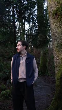 Γιλέκο ναυτικού στρωμένο πάνω από ένα ανοιχτό μπεζ τοπ, μαύρο παντελόνι, με ουδέτερη υφή υφάσματος. Περιβάλλεται από πράσινο και ψηλά δέντρα.