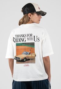 Femme portant un t-shirt blanc avec le texte « Merci d'avoir roulé avec nous » et une image d'une personne sortant d'une voiture décapotable beige près d'un court de tennis.