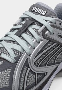 Detailný záber na sivú a svetlomodrú bežeckú topánku Puma s vrchnou časťou z priedušnej sieťoviny, detailnými prekrytiami a prekríženými šnúrkami s nápisom "Sky Jet".