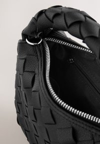 Sac à main noir à texture tissée, fermeture éclair et garnitures argentées. L'intérieur comprend une petite poche zippée contre une doublure en tissu.