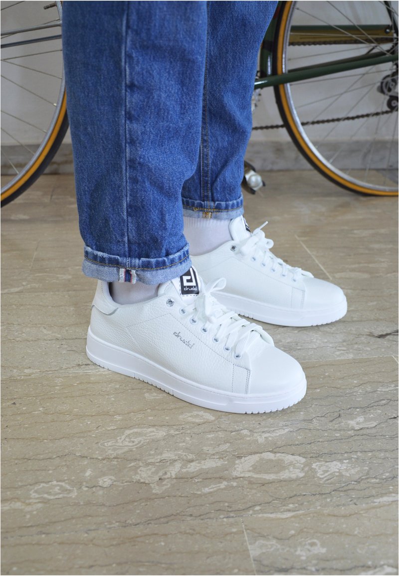 Sneakers in pelle bianca con finitura testurizzata, punta rotonda e suola piatta in gomma. Presentano lacci bianchi e una etichetta con il marchio sulla linguetta. Indossati con jeans blu.