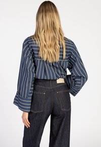 Blonde vrouw met golvend haar draagt een blauwe gestreepte blouse met lange mouwen, in haar hoge taille donkere spijkerbroek met achterzakken gestopt.