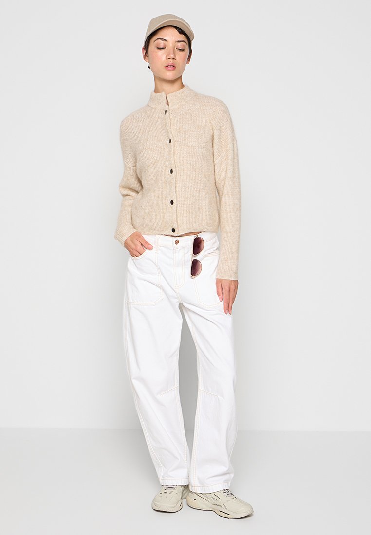 Beige gestrickte Strickjacke mit hohem Kragen und schwarzen Knöpfen, kombiniert mit weißen weiten Hosen und Turnschuhen. Eine Sonnenbrille hängt am Bund.