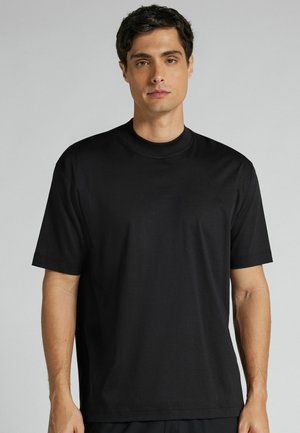 Mand med kort mørkt hår iført en almindelig sort T-shirt med korte ærmer og rund hals, står foran en lys grå baggrund.