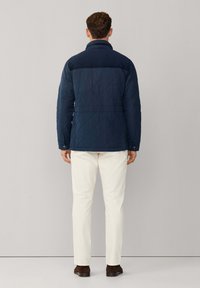 Marineblauwe gewatteerde jas met opstaande kraag, met een gladde textuur en tonale accenten op de schouders, gecombineerd met witte broek en bruine schoenen.