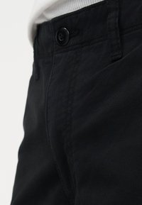 Κοντινή λήψη στη μέση μαύρου παντελονιού με μαύρο κουμπί κλεισμένο και μέρος λευκού πουκάμισου που έχει μαζευτεί μέσα.