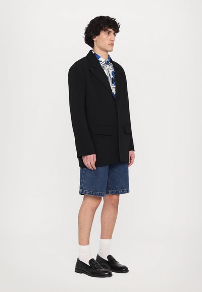 Black oversized blazer, denim shorts, patterned shirt, black loafers, and white socks. Features sharp lapels and two front pockets.