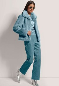 Faux-Fur-Jacke in Petrol mit Kragen, kombiniert mit gerippten petrolfarbenen Hosen und glitzernden silbernen High Heels. Das Modell trägt runde Sonnenbrillen.