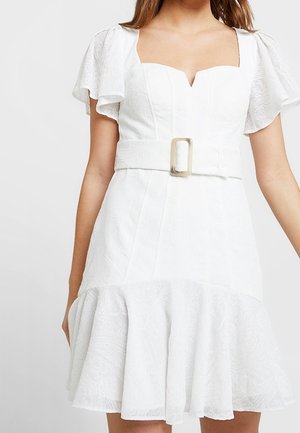 Valkoinen mekko, jossa on lyhyet, liehuvat hihat, teksturoitu kangas, tiivis yläosa ja levenevä, röyhelöreunus. Mukana on leveä vyö soljella.