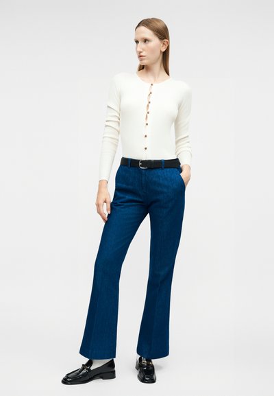 Γυναίκα που στέκεται με τα χέρια στις τσέπες, φοράει λευκό μακρυμάνικο πουκάμισο με κουμπιά, μπλε ψηλόμεσα παντελόνια με ζώνη και μαύρα μοκασίνια με λευκές κάλτσες.