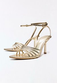 Sandale aurii metalice cu toc înalt, cu bretele subțiri la gleznă, multiple bretele în față și vârfuri deschise ascuțite pe un fundal alb.
