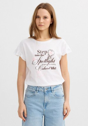 Joven mujer con cabello largo y castaño, que lleva una camiseta blanca con mangas con volantes y una cita impresa, y vaqueros de talle alto de color azul claro.