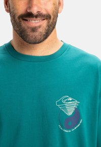 Camiseta de algodón color aguamarina con un escote redondeado, que presenta un diseño gráfico blanco que incluye una ola y un símbolo de yin-yang.