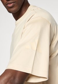 Béžové tričko s krátkym rukávom vyrobené z hladkej tkaniny, s tónovým logom s tromi pruhmi na hornej časti rukáva a klasickým okrúhlym výstrihom.