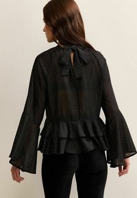 Donna che indossa una blusa nera trasparente con pois bianchi, maniche a campana, orlo peplum stratificato e un fiocco legato al colletto sul retro.