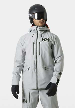 Uomo che indossa giacca da sci grigio chiaro e pantaloni con guanti neri, casco nero e occhiali da sci, volto leggermente rivolto a destra su sfondo neutro.