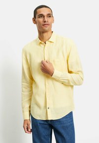 Helles gelbes Leinenhemd mit langen Ärmeln, spitzem Kragen, Knöpfen vorne und einer Brusttasche; kombiniert mit blauen Jeans.