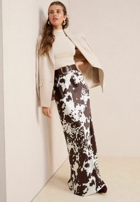 Jupe en satin marron et blanc à motif vache, longue, associée à un col roulé beige et un blazer clair, rehaussée par une ceinture marron foncé.