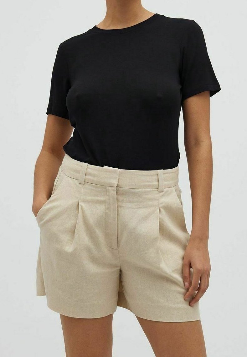 Femme portant un haut noir à manches courtes rentré dans un short beige taille haute avec plis et poches latérales, debout devant un fond uni.