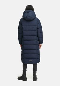 Casaca lungă puffer bleumarin cu glugă, secțiuni matlasate și un accent cu logo la tiv. Purtați cu pantaloni negri și cizme.