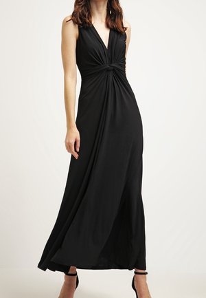 Femme portant une robe maxi noire sans manches avec un détail de nœud torsadé à la taille et des talons noirs à bride autour de la cheville.