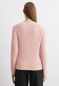 Femme aux longs cheveux raides portant un pull en tricot rose clair et un pantalon noir, se tenant face à un mur blanc.