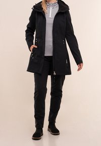 Vrouw in een lange zwarte jas, zwarte broek, zwarte laarzen en een wit-zwarte gestreepte zip-up top, staande op een lichte achtergrond.